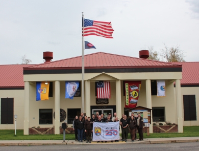 A group of people in brown holding a white “America 250” flag stand in front of a tan building with a red roof. Flags from USFWS, the Confederated Tribes and Bands of the Yakama Nation, the Betsy Ross flag, the Confederated Tribes of the Colville Reservation, and the Bureau of Reclamation flag fly behind. US flag and fish pennant fly above