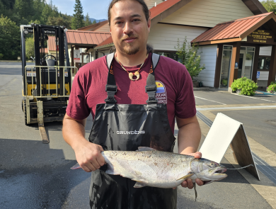 Addison Emerson holding an adult coho salmon at Quilcene National Fish Hatchery. Forklift, facility, and skyline in the background.