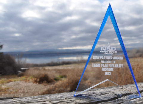 A translucent rectangular award sits in front of a lake and sagebrush habitat with an engraving that reads “Nampa School District New Partner Award Presented To Deer Flat National Wildlife Refuge 2023”