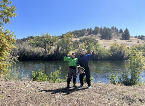 Two smiling biologists flex their muscles along a river bank