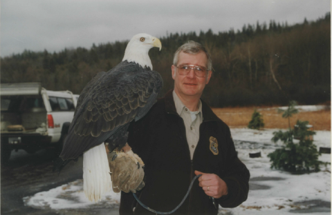 A bald eagle perches on the arm of a man in a Service jacket in a snowy forested outdoor setting