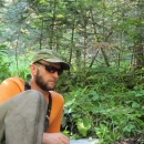 Fish Biologist, Brian Davis, collecting samples in the field. Brian is wearing an orange shirt, tan pants, tan hart and black sunglasses. He is reclining on the ground in a temperate rainforest setting with sampling equipment in front of him. 