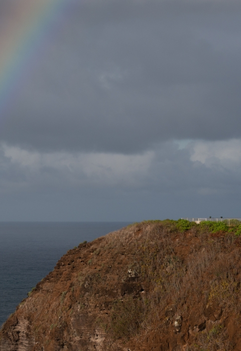 A rainbow arcs over the ocean and a lighthouse sitting on a rocky point. 