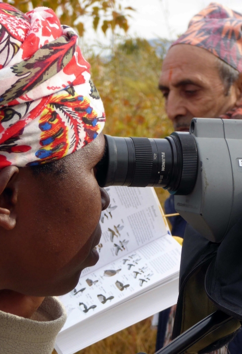 A woman looks through a spotting scope, and a man can be seen in the background behind the scope