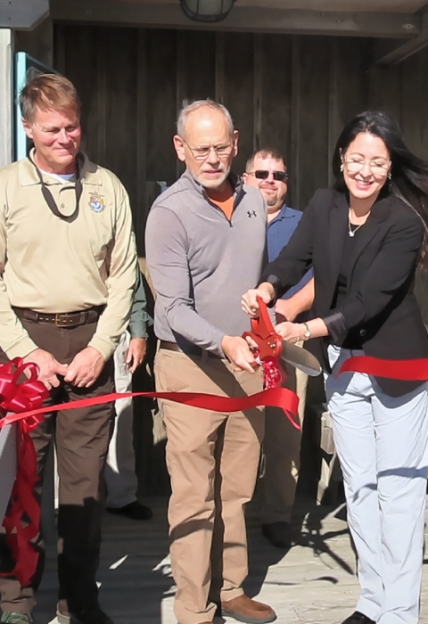 A group of five men and women cutting a ribbon with a pair of oversized scissors. There are a few onlookers in the background.