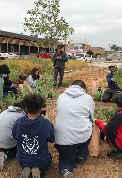 A group of students kneel next to each other in a semi circle as their teacher stands on the left end of the semi circle. Each student has a single potted plant in front of them. They are either looking at their plant or listening to their instructor who is also in a kneeling-sitting position holding out a green and shrubby potted plant.