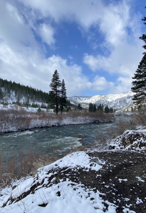 A landscape photo of a river flowing between mountains dusted with snow under a partly cloudy blue sky.