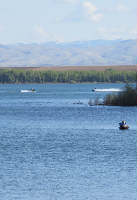 A kayaker is fishing and two people are riding personal watercraft on a calm lake. There are trees on a point in the foreground and on the distant shoreline, as well as mountains in the distance. 