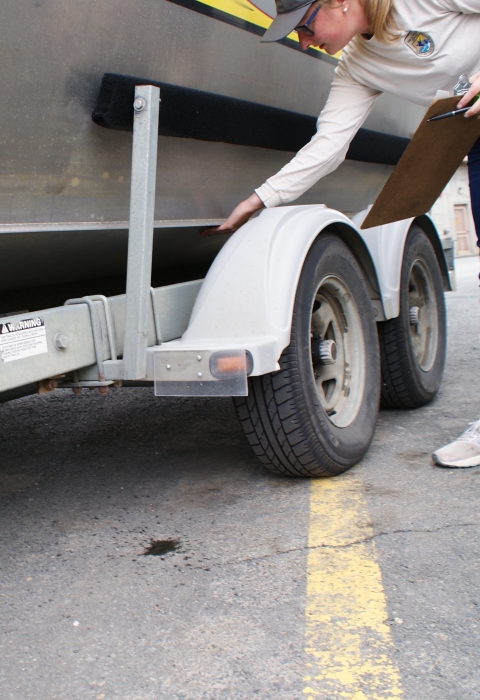 Watercraft inspector feels underside of trailer for aquatic invasive species while holding a clipboard.