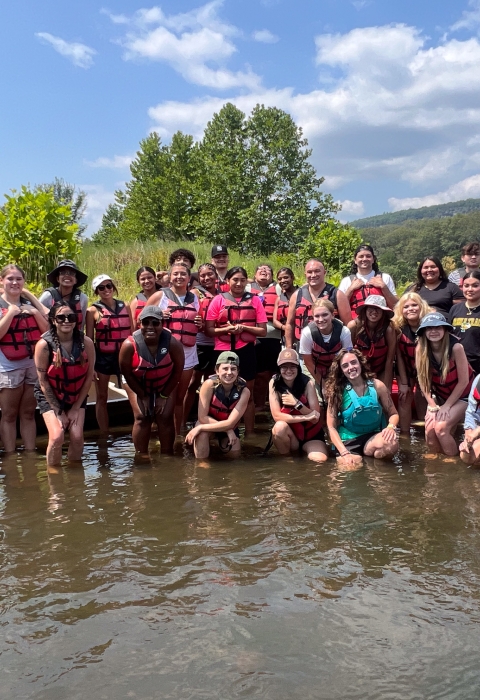 Many smiling people wearing life jackets standing in shallow water with canoes and a forested horizon.