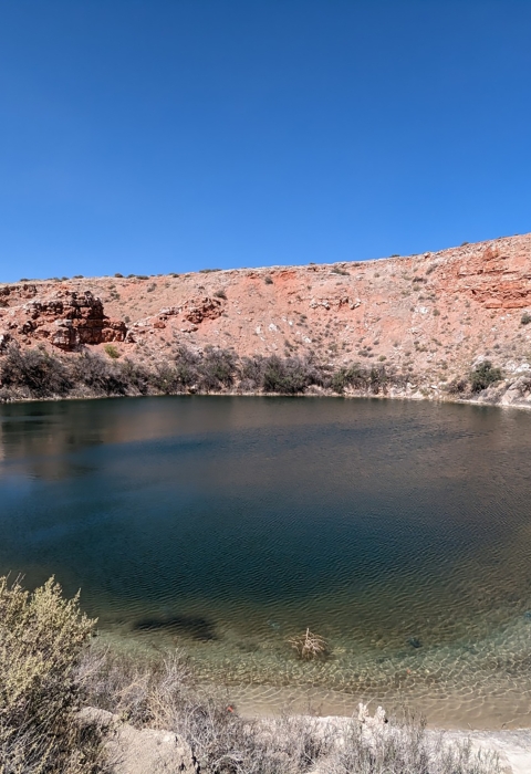 A shallow pool is encircled by rocky red bluffs and sagebrush on a bright sunny day.