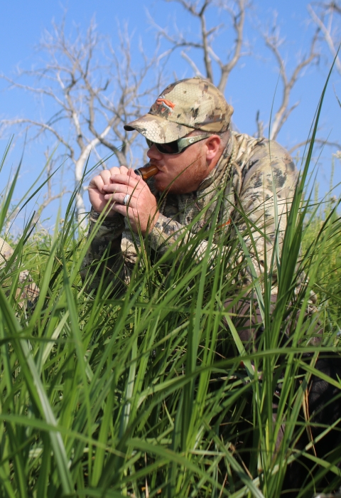 Two men dressed in camouflage sitting in grass. One man is holding a duck call to has mouth.