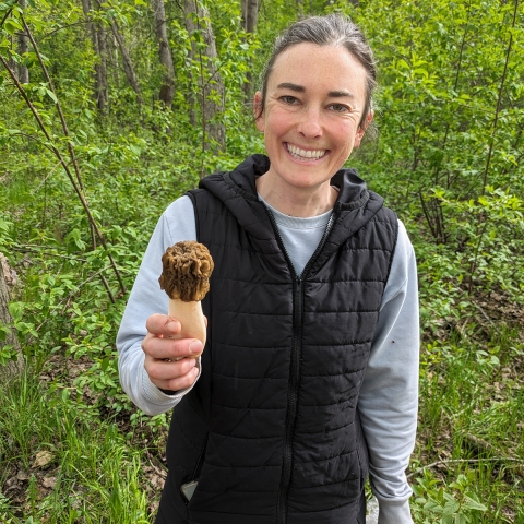 In a forest a person smiles and holds a mushroom in their hand. 