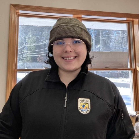 A woman in a green winter hat and a black jacket with the U.S. Fish and Wildlife Service logo on the chest. She wears large square glasses and stands in front of a window. 