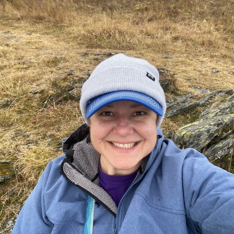 A person wearing a visored hat under a knit cap and a rain jacket takes a selfie with tundra and rocks behind them.