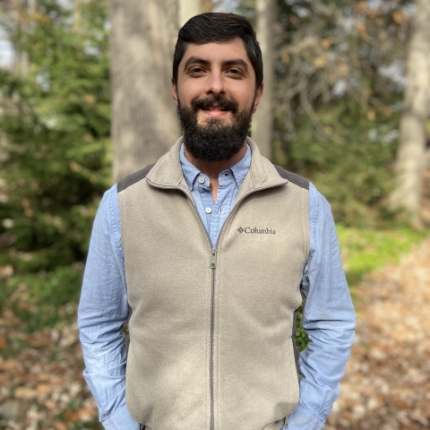 Alex stands smiling wiht hands in his pockets. He is wearing a tan fleece vest and a light blue button down shirt