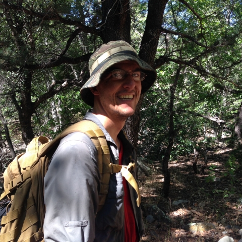 A man with a hat and backpack on in a forest smiling