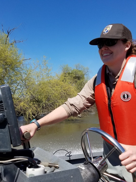 A woman drives a boat wearing a life vest and Fish and Wildlife Service hat