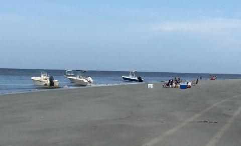Three boats sit at the shoreline while people sit on the beach nearby.