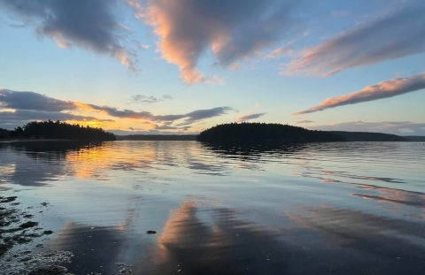 Streaks of brightly lit clouds at sunrise reflect on a body of water with distant islands