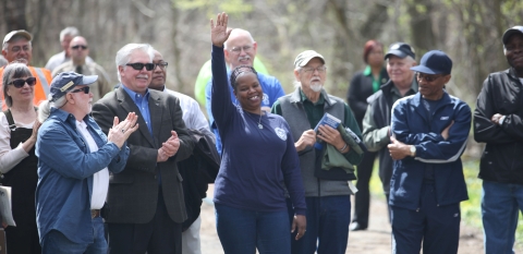 A smiling woman in the center of a group raises her hand while others around her applaud.