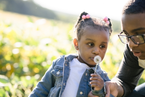A young girl blows on a puff of dandelion seeds that she picked from the side of the road. She is with a boy, probably her older brother, enjoying the sunshine at the edge of a field. 