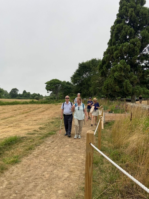 A man with a white cane and sunglasses walks alongside a woman in a green tee shirt and khaki pants. They are both on a trail nearby an open field. Behind them other visitors walk the trail. 