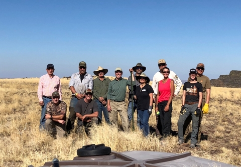 A group of people stand together in front of a water guzzler, surrounded by desert grass with cliff faces in the far distance.