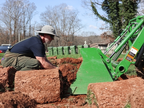 Person crouching in front of a tractor, lifting rolls of sod into the tracot bucket