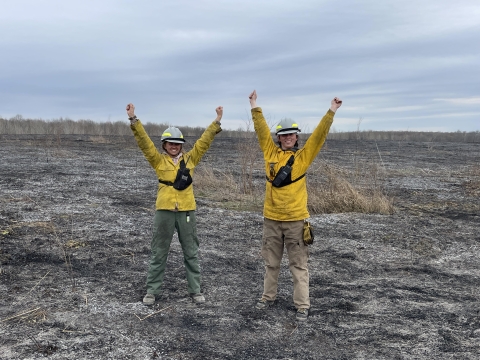 Two wildland firefighters stand in the black after a prescribed burn smiling with arms raised triumphantly
