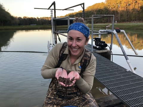 Jessica Radich holds freshwater mussels while wearing waders and standing in shallow water in front of a dock.