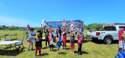 a group of children hold fishing rod packages in a green space with a U.S. Fish and WIldlife Service vehicle and trailer behind them.
