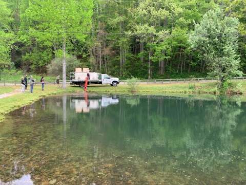 A large green pond is pictured. At the opposite end of the pond sits a fish stocking truck equipped with tanks and an orange hose attached that runs into the pond.