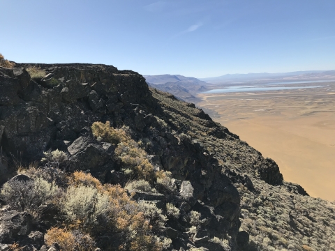 Rocky cliff faces covered in sagebrush rise above a valley floor full of fields and wetlands.