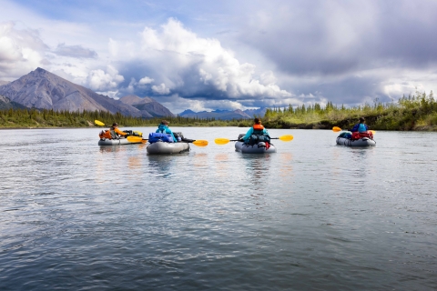 View of four people floating a river with mountains and a cloudy sky in the background. 