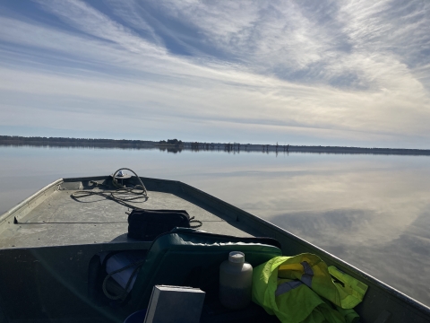 The prow of a boat looks out over a glassy lake surface reflecting cloudy sky and forests on the horizon.