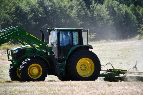 A green and yellow tractor tows a mowing attachment across a dusty field of grass with evergreens in the background.