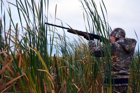 Female standing in tall grass wearing camouflage hunting outfit holding a rifle and aiming to the left.