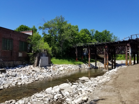 A river flows from upper right to lower left, with part of a red brick building and green trees on the left bank and white rocks and soil on the other. An old railroad bridge is in the background.