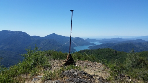 A tall pole on the top of a mountain with a microphone at the top. This pole is secured with anchors to the rocks below. In the background is a bright blue river flowing through the valley of mountains. 