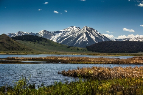 A snowcapped mountain sits in the background under a blue sky in front of green hills and a lake