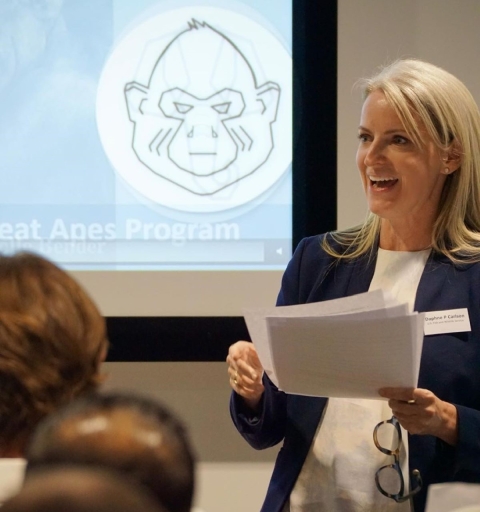 A closeup of a woman presenting to a crowd. She has shoulder-length blonde hair, wearing a navy blazer and a white shirt, and his holding papers and a pair of glasses. 