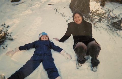 A woman, to the right of the frame, sits in a patch of snow and smiles up at the camera while her daughter, to the left and wearing a blue beanie and white gloves, makes a snow angel.