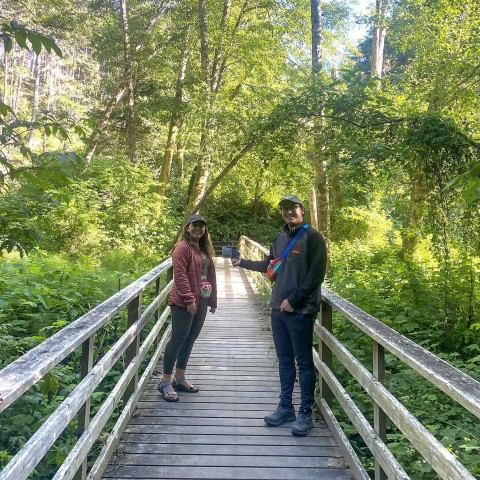 Two people stand on either side of a wooden bridge, framed by green vegetation in the background, and smile as they face the camera.