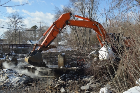 The orange arm of a construction excavator reaches into a river to break apart a dam