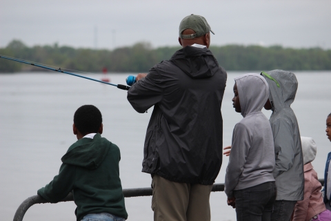 A man stands on the waterfront with a fishing rod surrounded by kids