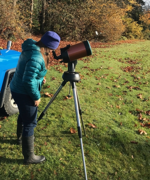 Jen Johnson looking through a telescope with a solar scope attached. She is wearing a green jacket, jeans, and boots and standing on grass with trees in the background.