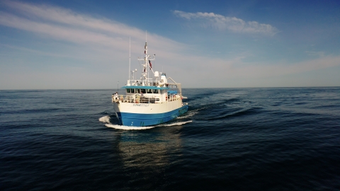 The Research Vessel Connecticut, a 90' steel blue and white ship with a tall mast and various antennas, is traveling through the water with no land in sight. The horizon stretches out far in the distance. The sky is blue, with white clouds.