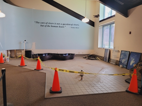 A large room showing the floor which is partly tile and partly carpeted. Sitting on the floor also are orange cones with hazard tape connecting the cones. This is being done to keep visitors out of a construction area.