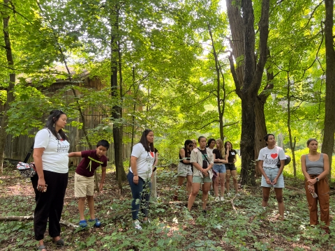 People wearing "I Heart Lenapehoking" shirts gather in a forested area under tall tree cover.